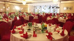 Une salle de banquet avec des tables rondes dressées pour un événement formel, agrémentée de chaises rouges, de nappes blanches et de compositions florales.