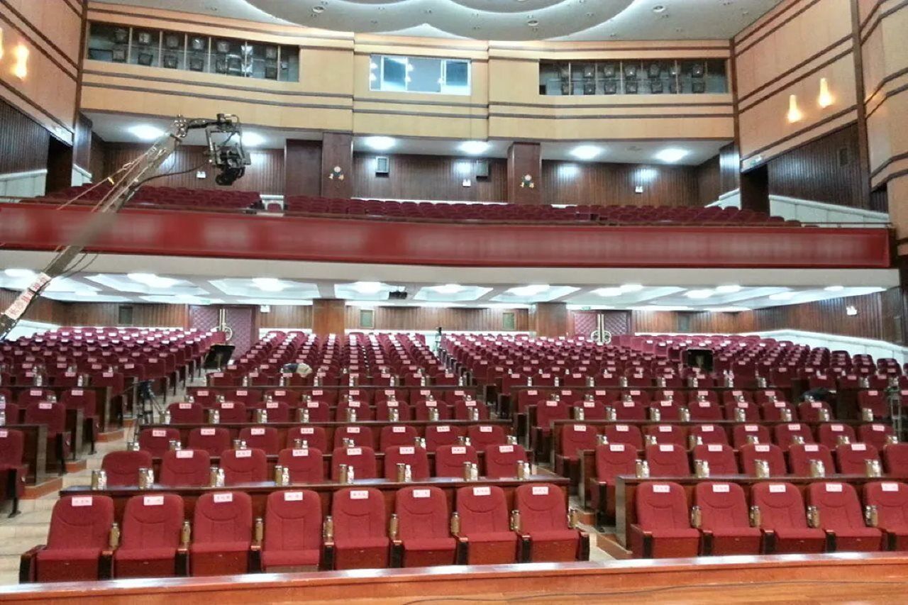 Un grand auditorium avec des rangées de sièges rouges, une scène équipée d’une caméra et une galerie avec des fenêtres.