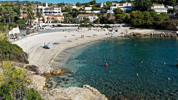 A coastal beach with clear waters, a sandy shore, and buildings in the background.