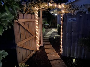 A wooden gate with a white archway adorned with string lights, leading to a garden area with a paved path and a wooden shed. - Your Wellington Getaway (Wellington)