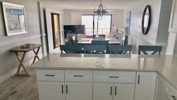 A modern kitchen with white cabinetry, a central island, and a dining area with blue chairs. - Ocean Front Luxury #801 (Cape Canaveral)