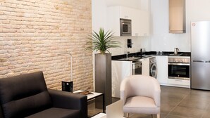 Una sala de estar moderna con un sofá negro, un sillón color beige y una planta alta. Un ordenador portátil está sobre la mesa de centro. La habitación tiene una pared de ladrillo y una cocina al fondo con electrodomésticos de acero inoxidable.