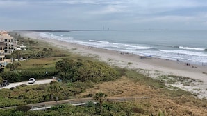 Una playa con vista al océano y un puente lejano. Hay edificios a lo largo de la costa y un estacionamiento.