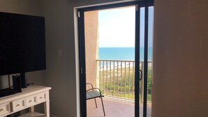 Una habitación con un televisor sobre un mueble blanco, una puerta corrediza de vidrio que da a un balcón con vista a la playa y al océano.