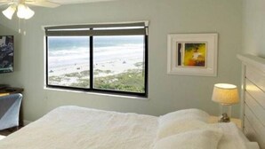 Un dormitorio con una cama grande, una ventana con vista a la playa, un cuadro en la pared y una lámpara en la mesita de noche.