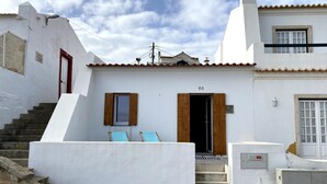 A white building with a number '50' above a wooden door, a small staircase leading to a balcony with two blue chairs, and a stone-paved ground. - Baleal Waterfront (Ferrel)