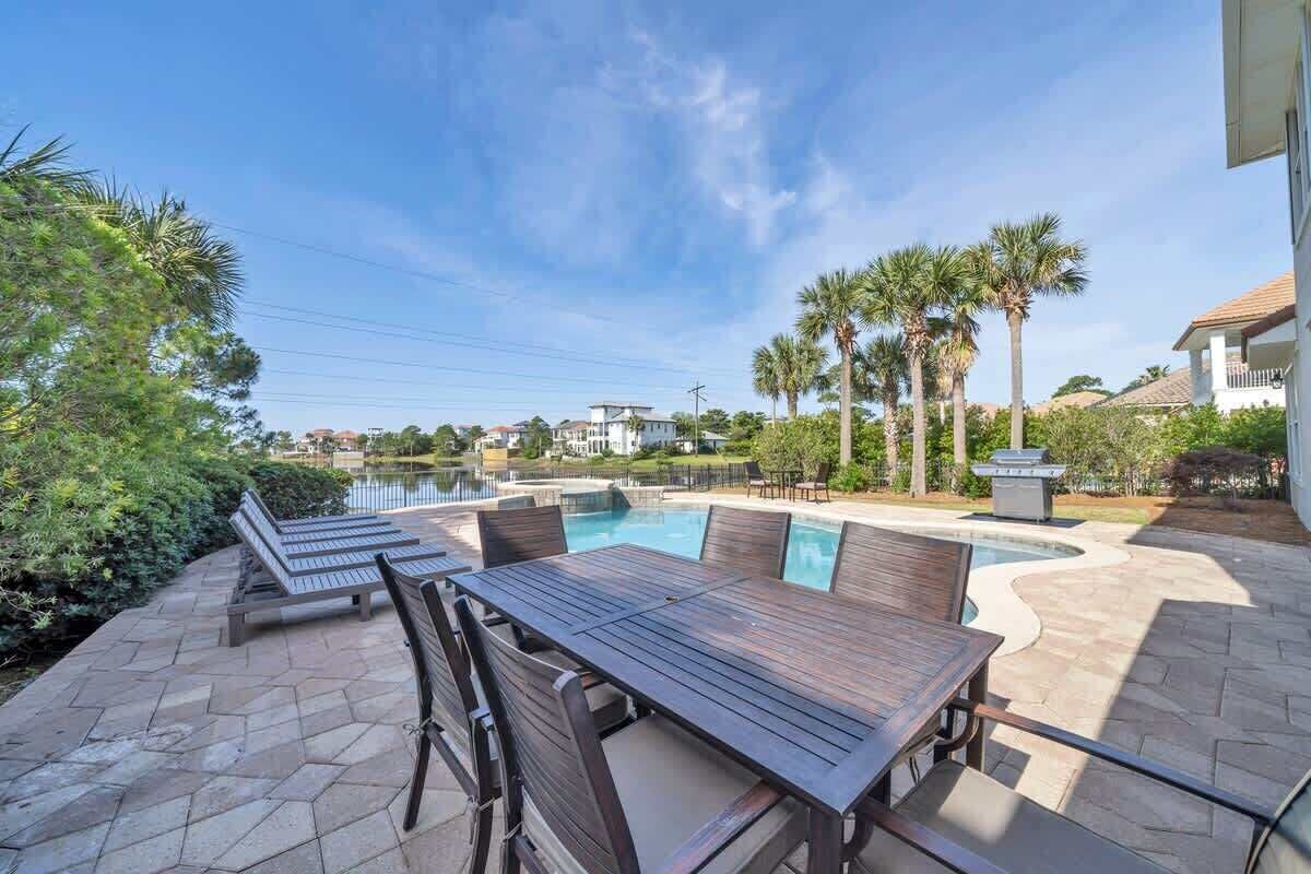 Un área de piscina con una mesa y sillas de madera, rodeada de palmeras y vista a un lago.