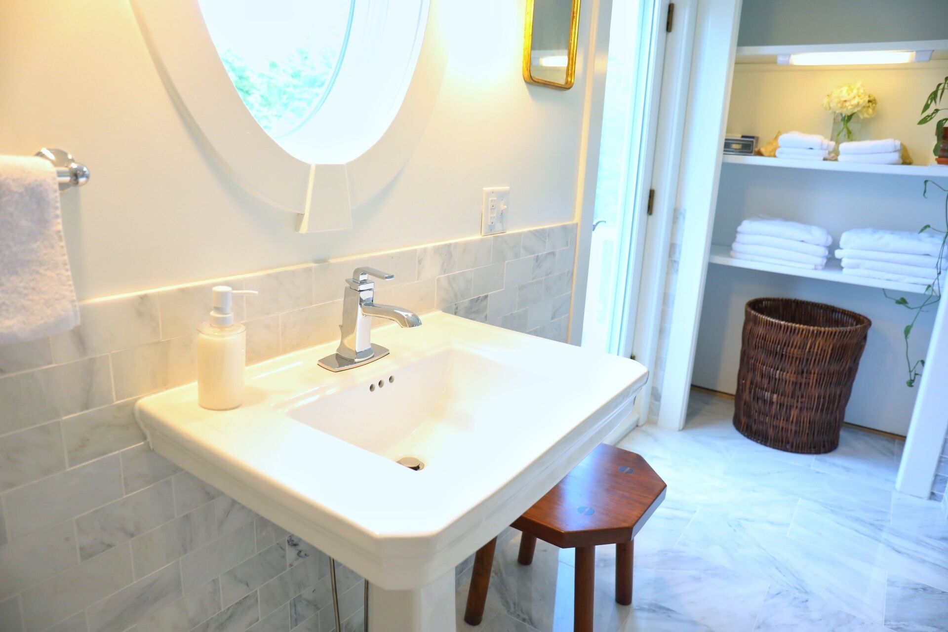 A bathroom with a white sink, a mirror, and a wooden stool. There is a basket and a shelf with towels in the background.