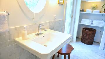 A bathroom with a white sink, a mirror, and a wooden stool. There is a basket and a shelf with towels in the background.