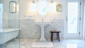 Ein Badezimmer mit Glasdusche, Marmorboden, einem weißen Waschtisch und einem kleinen Holzschemel.