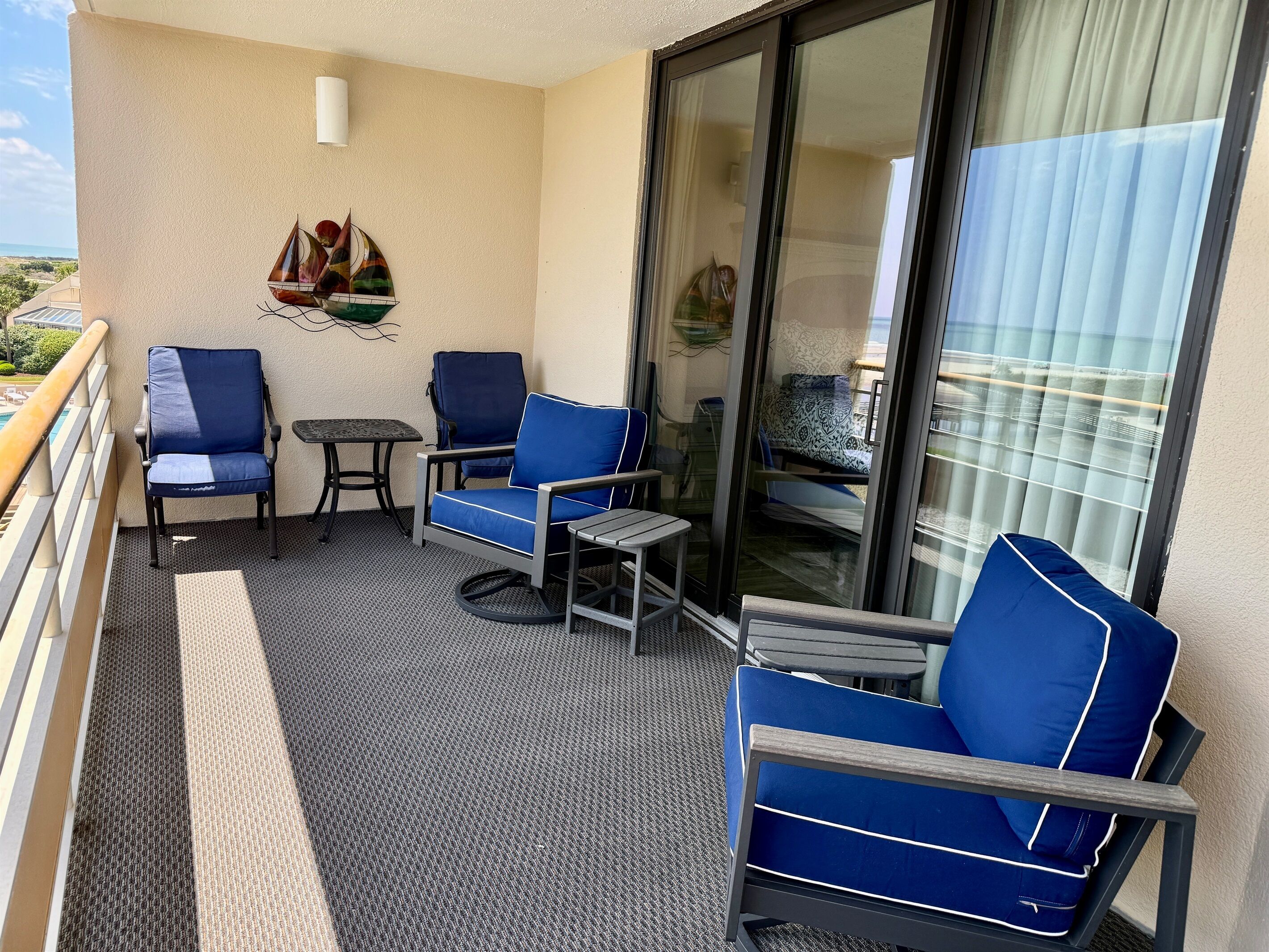 A balcony with two blue chairs, a small table, and a glass door leading to a room with a patterned bedspread.