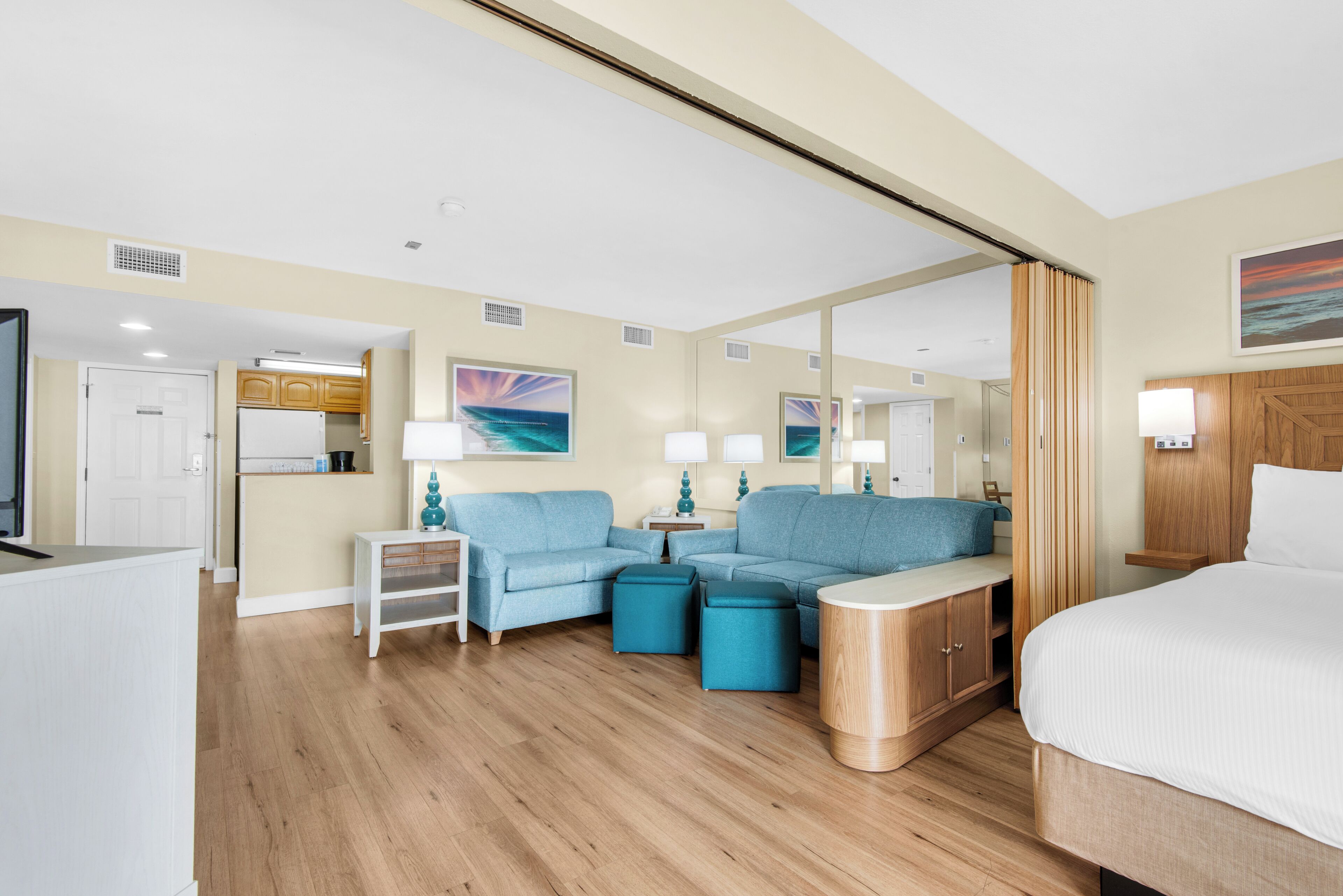 A hotel room with a bed, a sofa, a coffee table, a side table, a mirror, and a view of the kitchen.
