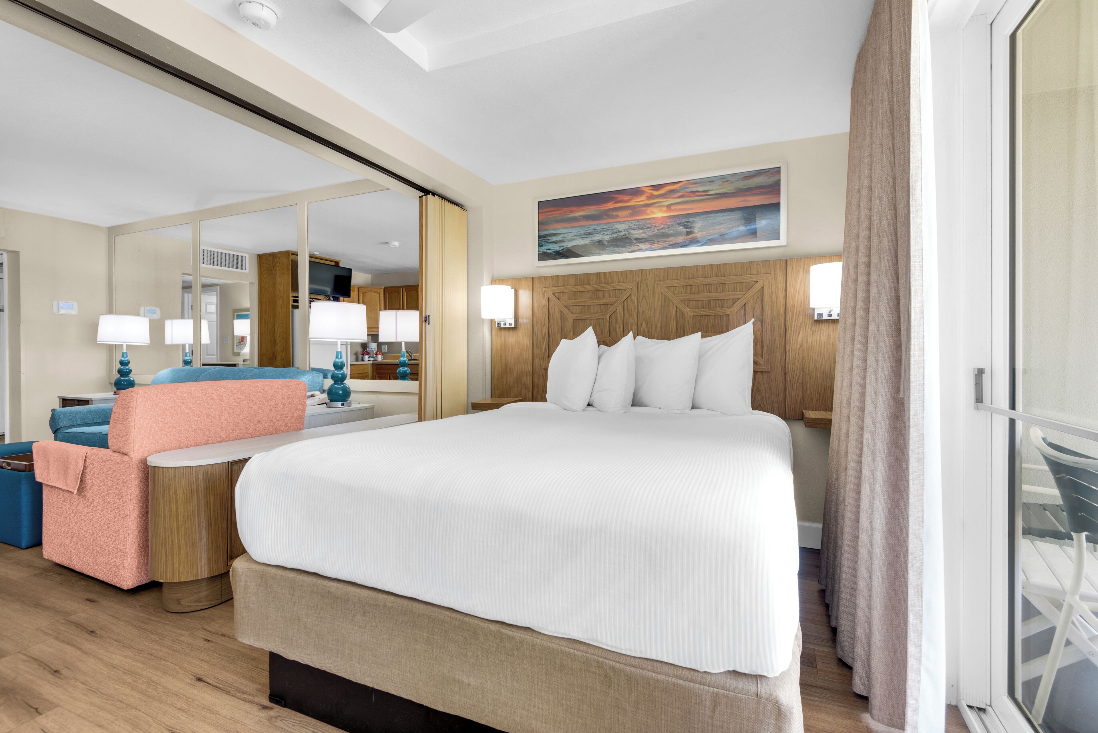 A hotel room with a large bed, a seating area with a chair and a table, a mirror, a painting on the wall, and a view through a window.