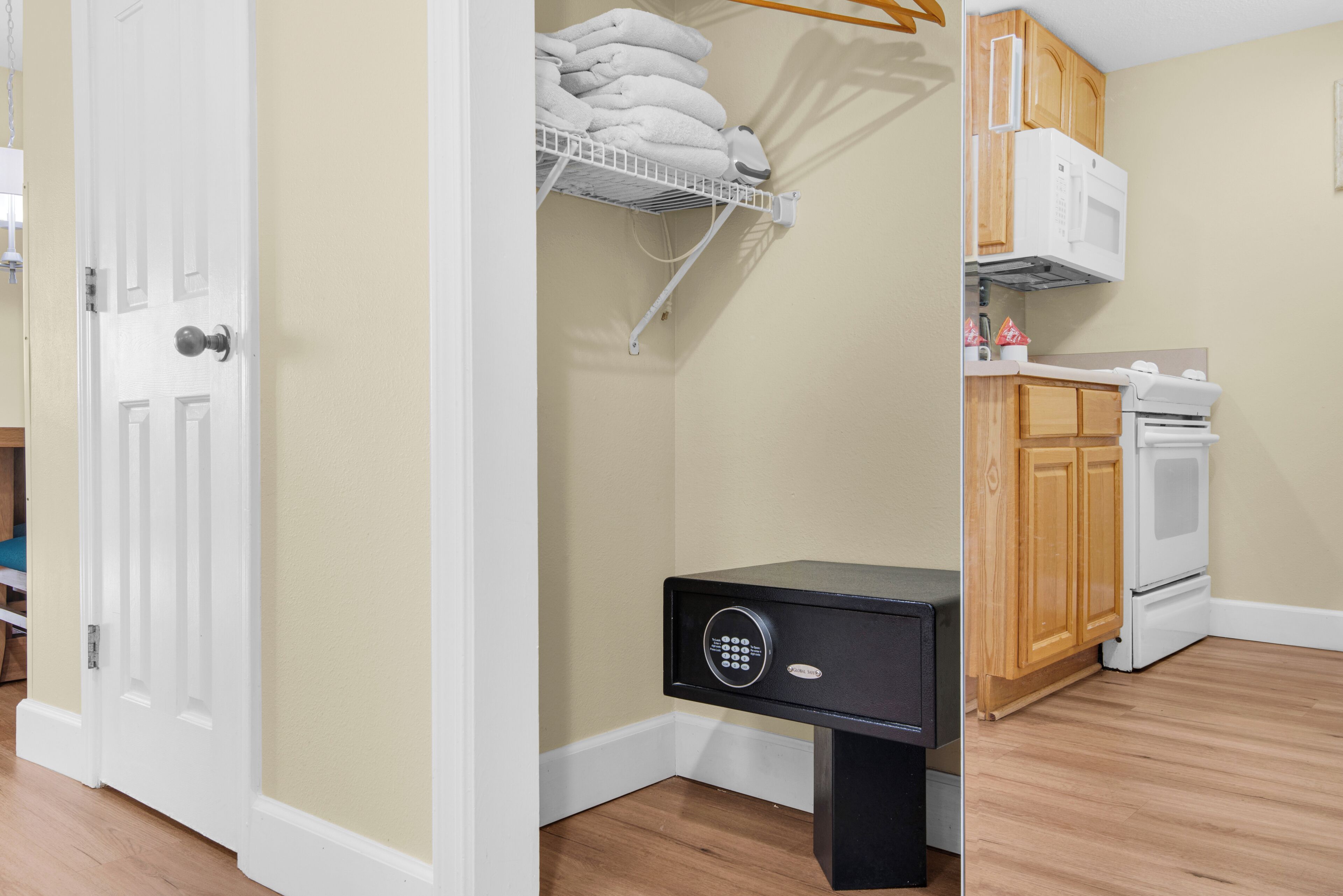 Una habitación con una puerta blanca, una caja fuerte negra, un microondas blanco, armarios de madera y un perchero blanco.