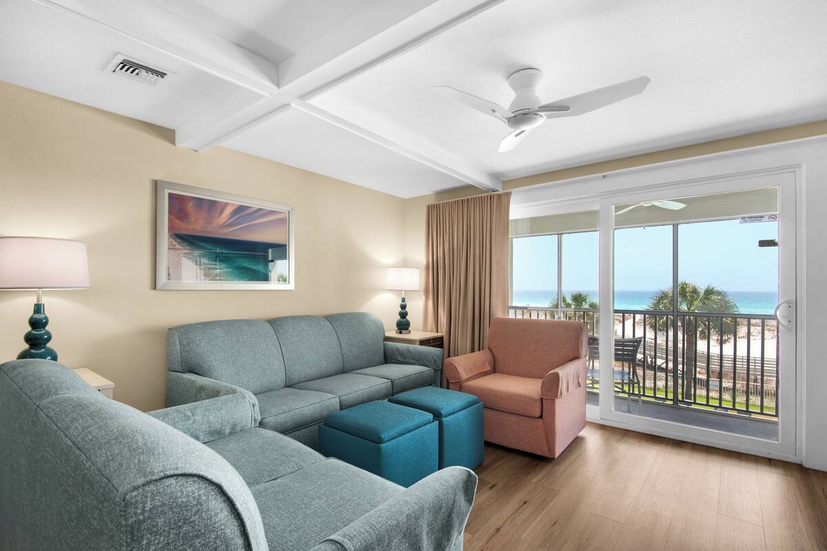 A living room with a sofa, ottoman, and armchair, a ceiling fan, a painting, and a balcony with a view of the beach.