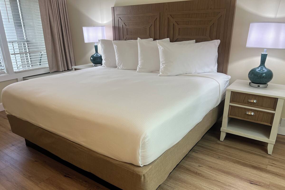 A large bed with white linens, a wooden headboard, two bedside tables with lamps, and a wooden floor.