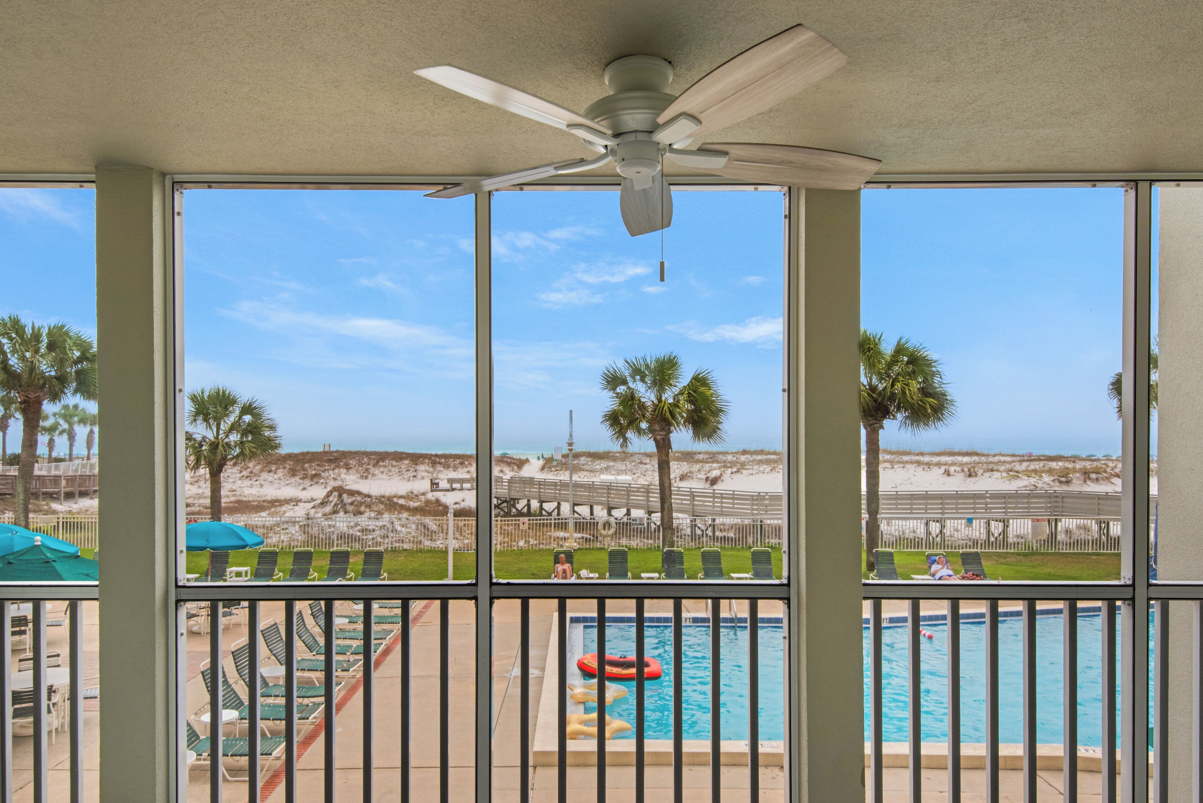 A pool area with a view of the beach, a ceiling fan, and lounge chairs.