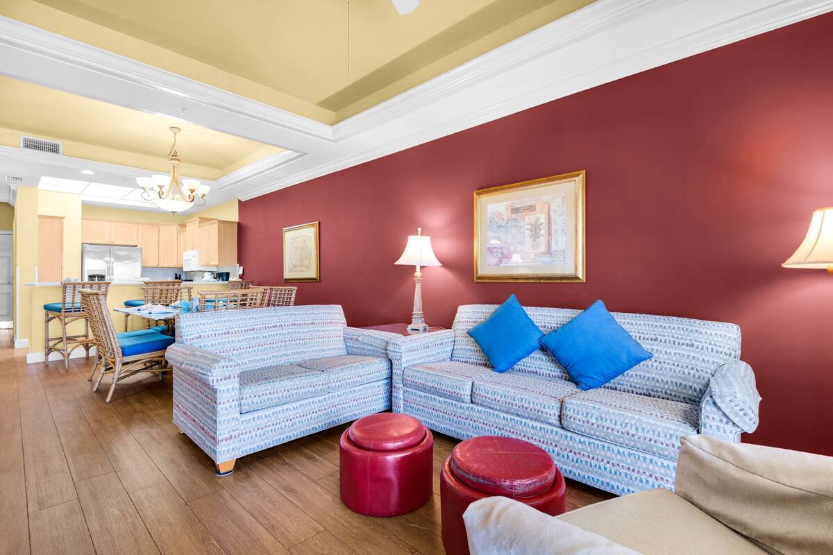 A living room with a patterned sofa, red ottomans, a dining area with wicker chairs, and a kitchen in the background.
