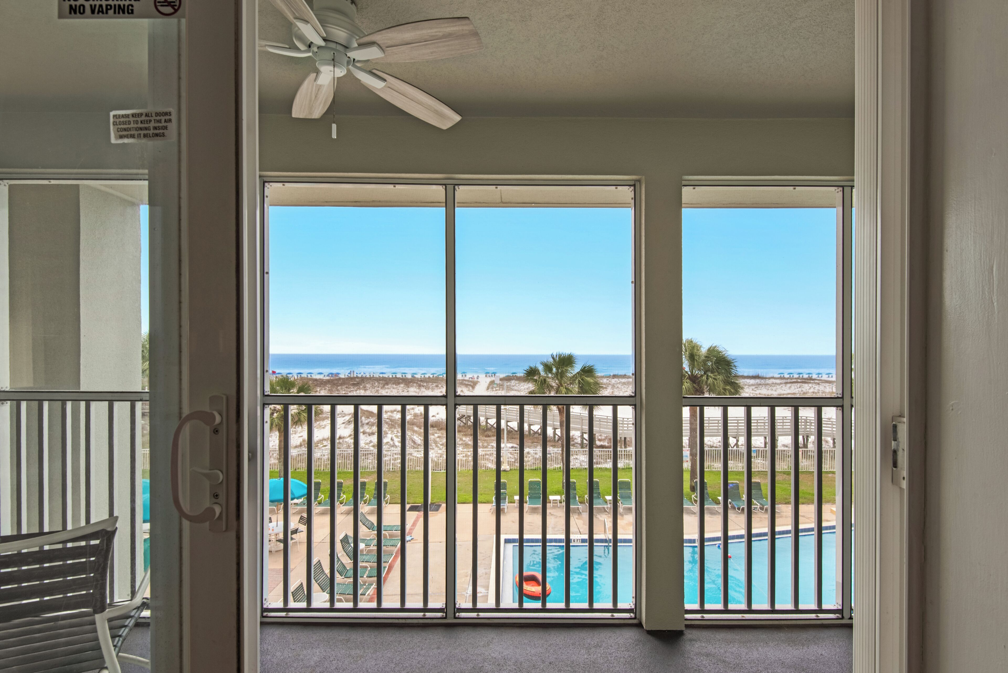 Un balcon avec vue sur la plage et la piscine, un ventilateur de plafond et un panneau indiquant qu’il est interdit de vapoter.