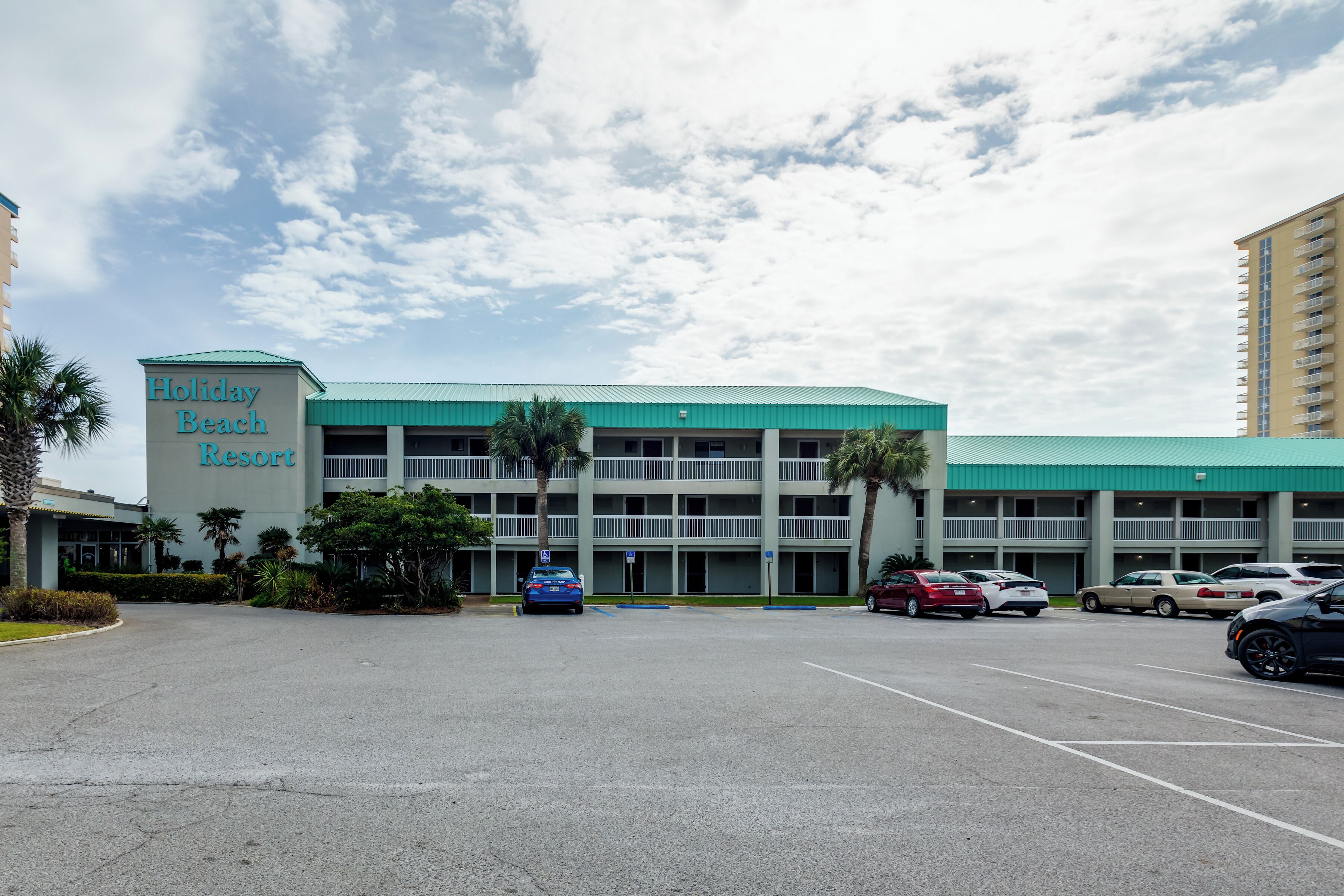 A Holiday Beach Resort with a parking lot, multiple floors, and a green roof.