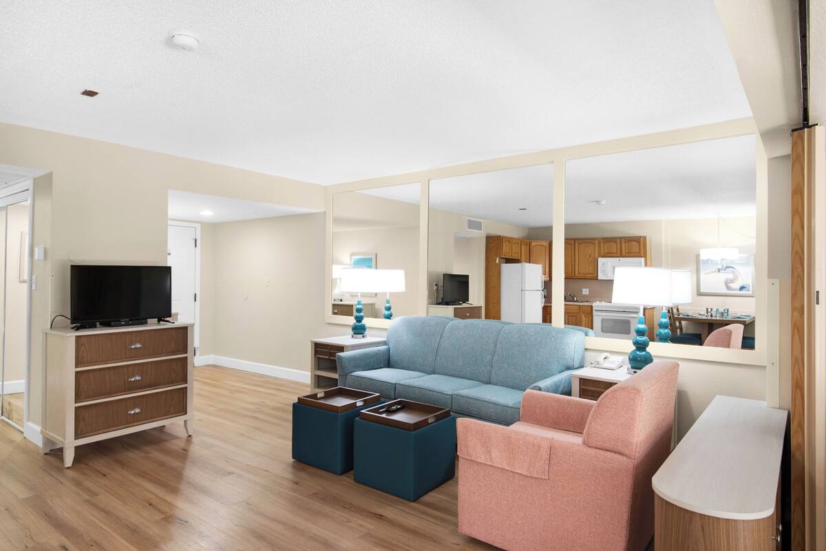 A living room with a sofa, ottomans, a television, and a mirror reflecting the kitchen area.