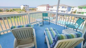 A balcony with white chairs and a small table, overlooking a pool and beach houses. - Cozy 2BR condo at Holden Beach, perfect for a family getaway! (Holden Beach)