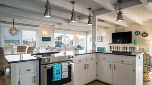 Une cuisine équipée d’une cuisinière, d’un four et de meubles blancs. On y trouve des luminaires suspendus, un téléviseur et une vue sur l’extérieur.