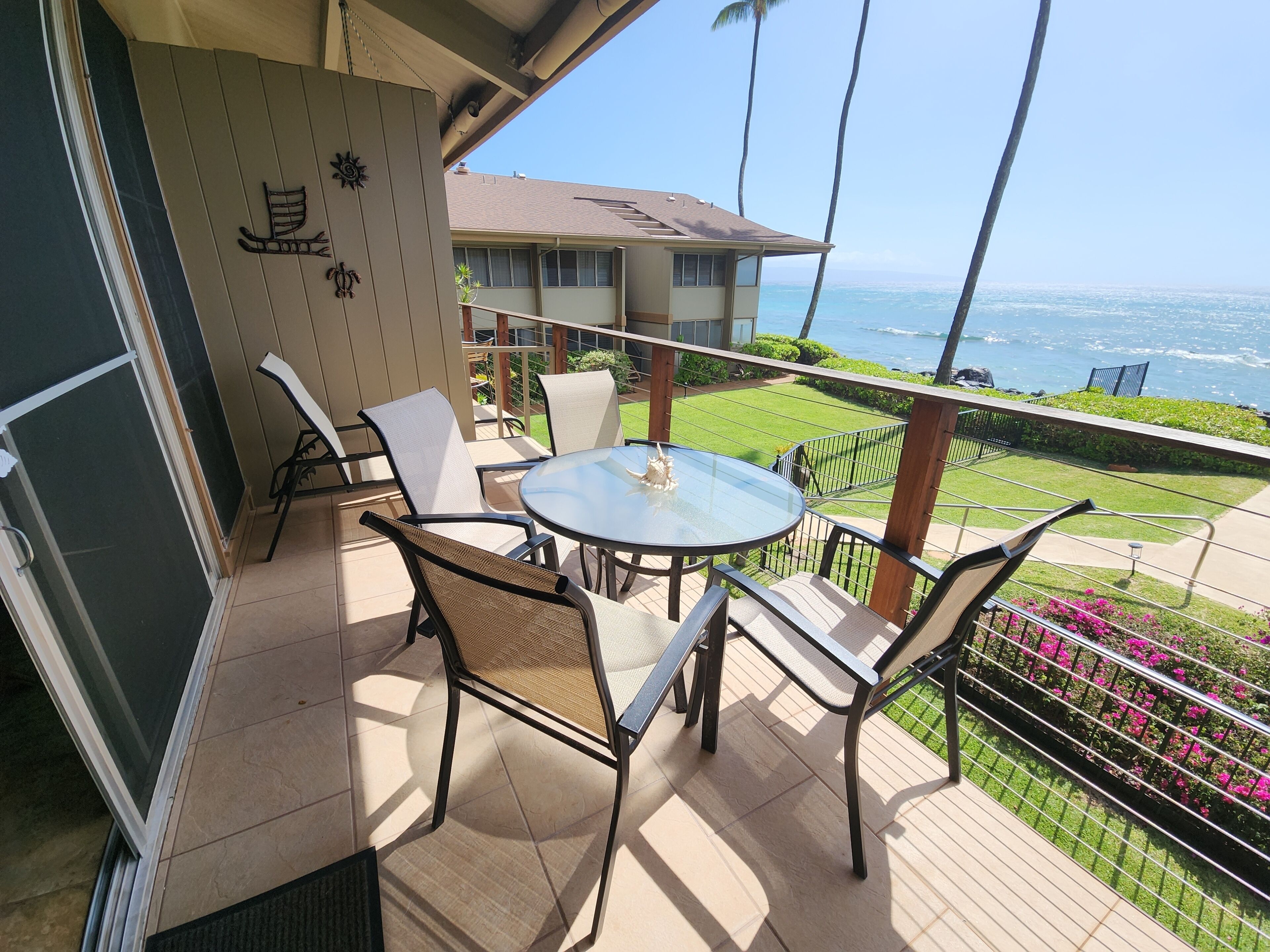 A balcony with a glass table and chairs, overlooking a green lawn and a view of the ocean.