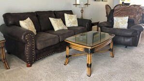 A living room with a dark brown leather sofa, a glass coffee table, and a patterned armchair. There are three framed pictures on the wall and a lamp on a side table.