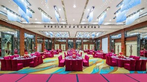 A large banquet hall with multiple round tables, each set with red chairs and white tablecloths, and a colorful carpet. - Golden Bay International Hotel (Meizhou)