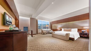 A hotel room with a large bed, a desk with a computer, a chair, and a view of the city through the window. - Golden Bay International Hotel (Meizhou)