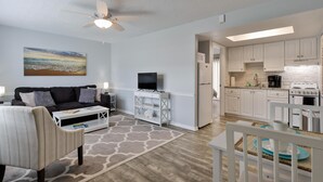 A modern living room with a sofa, TV, and coffee table, connected to a kitchen with white cabinets and appliances. - Coastal Waters 214, 2nd floor 1 bedroom ocean view condo (New Smyrna Beach)