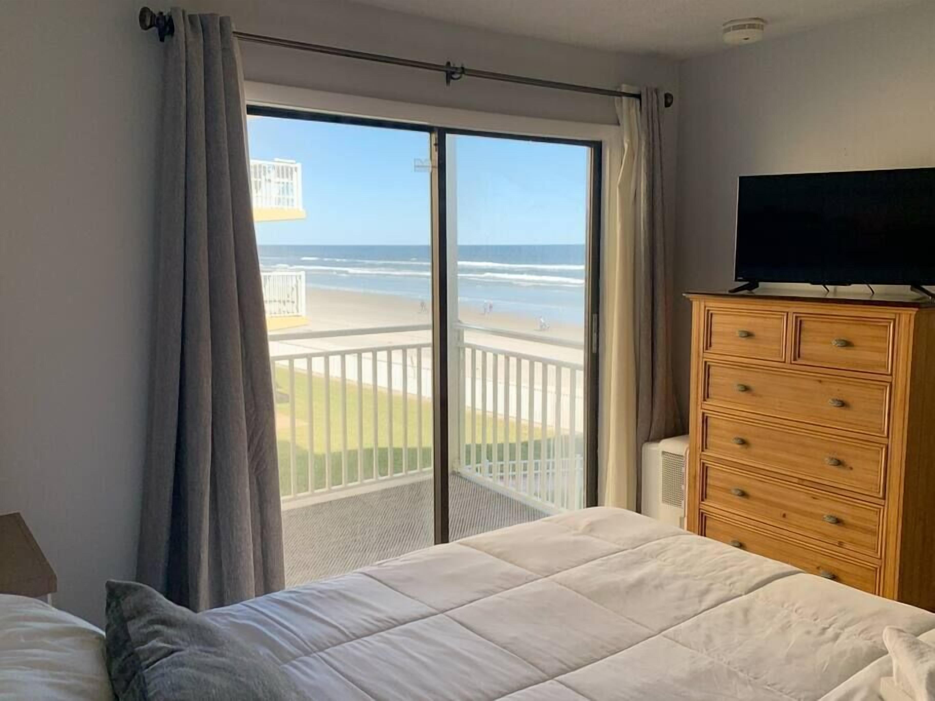 A bedroom with a bed, a wooden dresser, and a television. A balcony with a view of the beach is visible through the window.