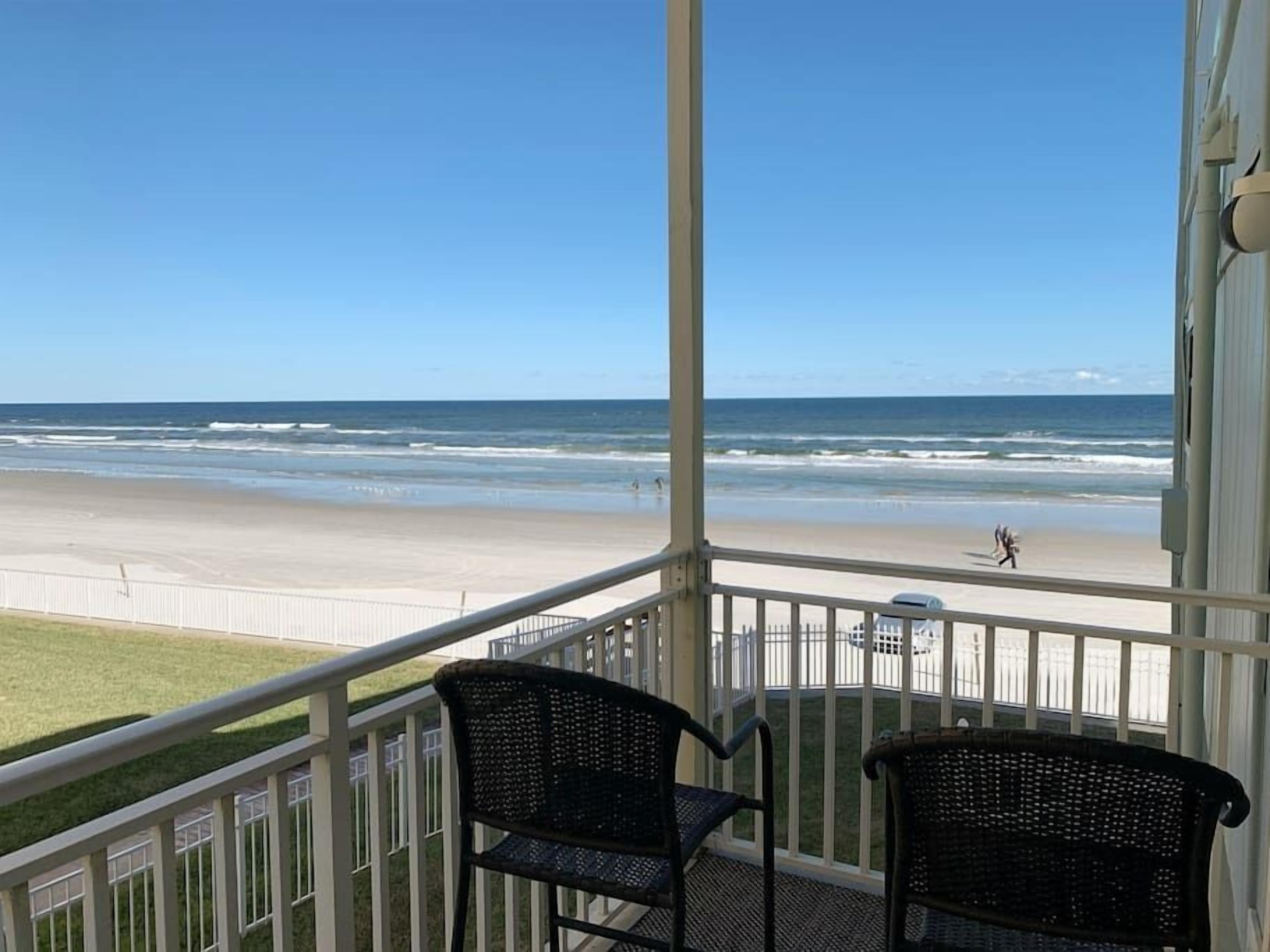 A balcony with two wicker chairs overlooking a beach with waves and a person walking.
