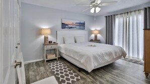 A bedroom with a bed, two bedside tables with lamps, a ceiling fan, a painting on the wall, and a view of the outdoors through curtains. - Coastal Waters 214, 2nd floor 1 bedroom ocean view condo (New Smyrna Beach)