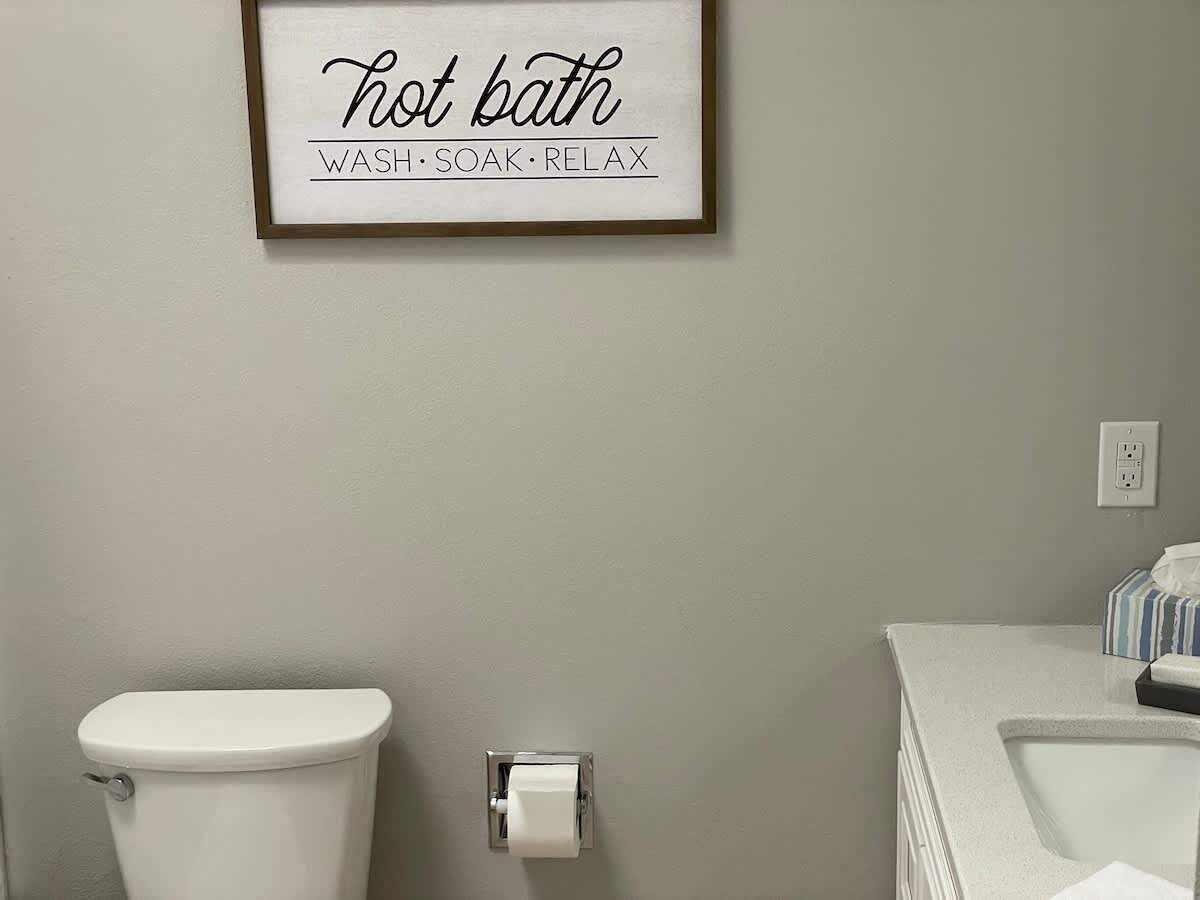 Un baño con inodoro, un rollo de papel higiénico y un cartel enmarcado sobre el inodoro.
