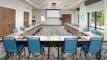 A conference room with a long table, blue chairs, a projector screen, and a view of the outside.