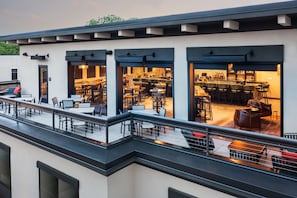 A modern outdoor dining area with multiple tables and chairs, a bar with stools, and a covered seating area with a view of the interior.