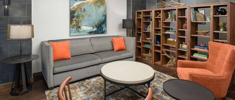 A modern living room with a grey sofa, orange chair, and round tables. A large abstract painting and a wooden bookshelf with decorative items are also visible.
