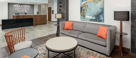Eine moderne Lobby mit einer Rezeption, einem grauen Sofa mit orangefarbenen Kissen, einem runden Couchtisch und einem gemusterten Teppich.