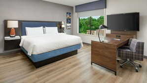 A modern hotel room with a large bed, a desk with a flat-screen TV, and a view of greenery outside.