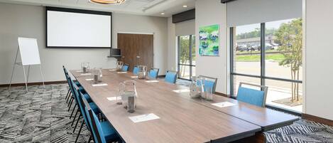 A conference room with a long table, blue chairs, a whiteboard, and a projector screen.