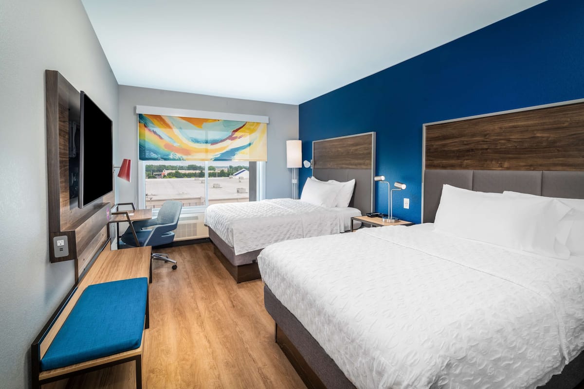 Habitación de hotel con dos camas, un escritorio, una silla, un televisor y un ventanal con vistas.