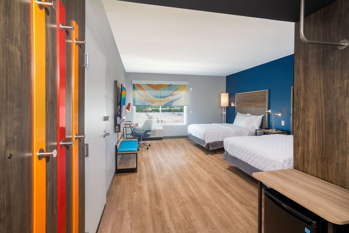Habitación de hotel con dos camas, un escritorio, una silla, una ventana con vistas y un cuadro abstracto y colorido en la pared.