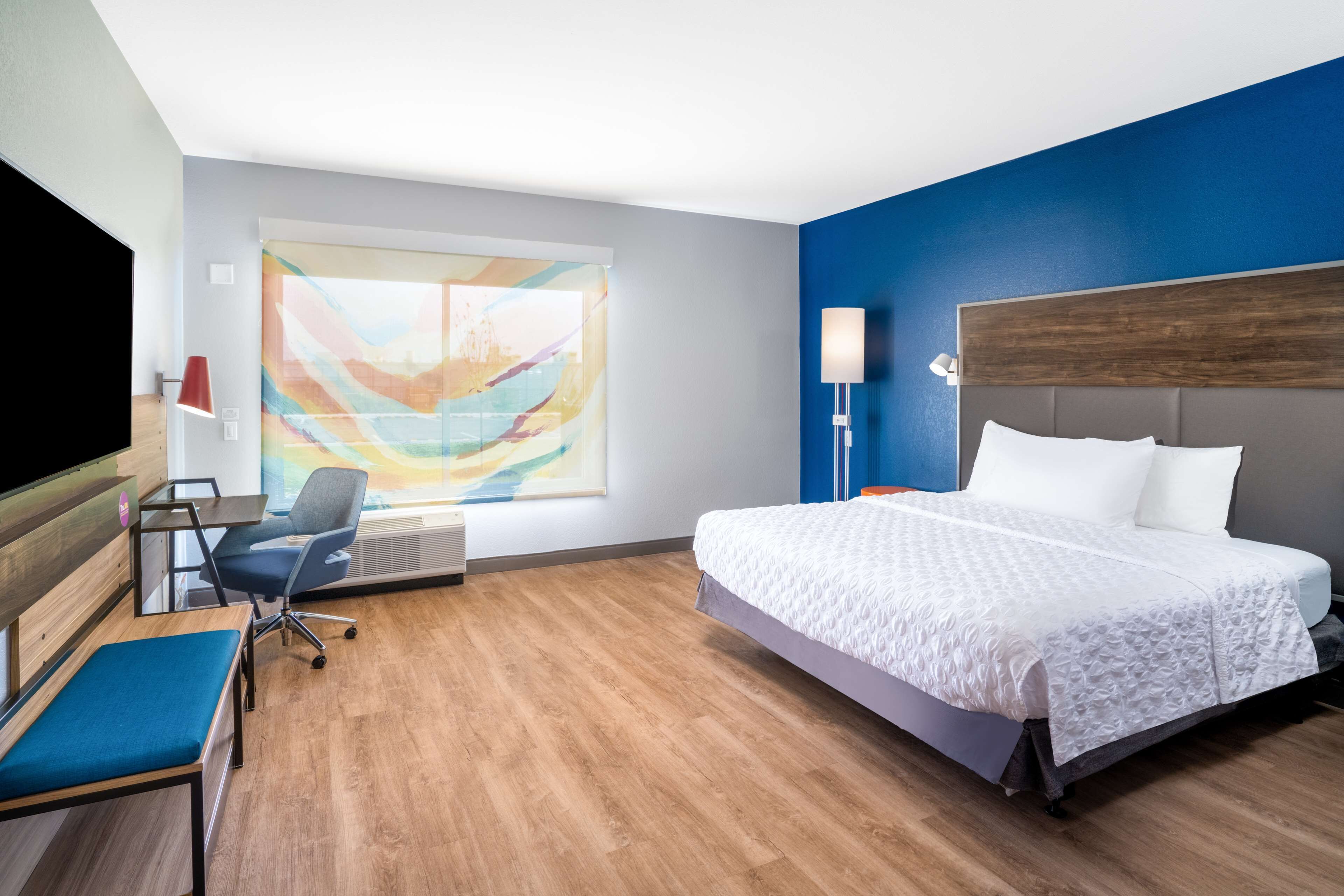 A hotel room with a large bed, a desk with a chair, a blue sofa, and a large window with a colorful abstract painting.