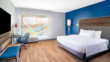 A hotel room with a large bed, a desk with a chair, a blue sofa, and a large window with a colorful abstract painting.