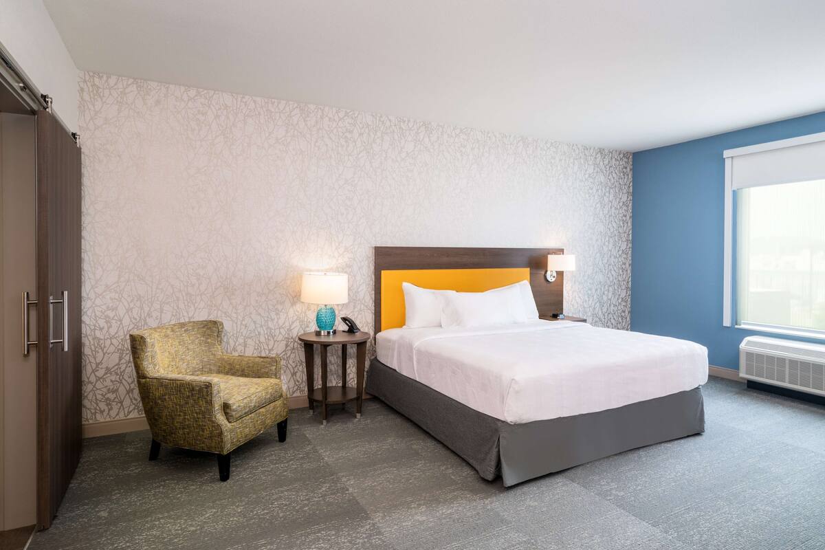 Una habitación de hotel con una cama, una silla, una mesita de noche con lámpara y una ventana.
