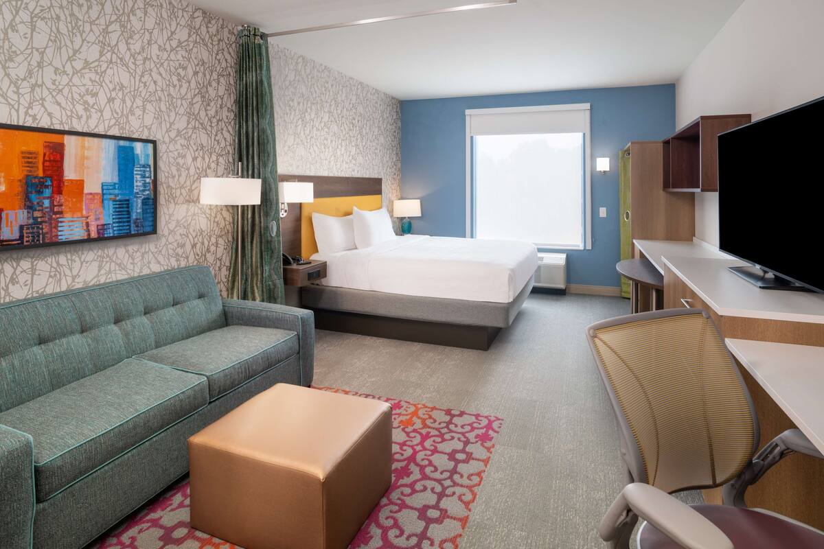 Una habitación de hotel con cama, sofá, escritorio, televisión y un cuadro de paisaje urbano en la pared.