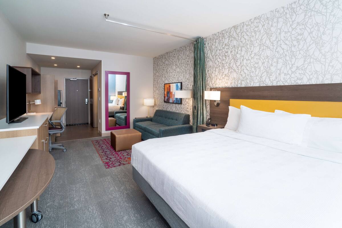 Habitación de hotel con una cama grande, un escritorio con una computadora, una silla y una mesita. Hay un espejo, una lámpara y un cuadro en la pared.