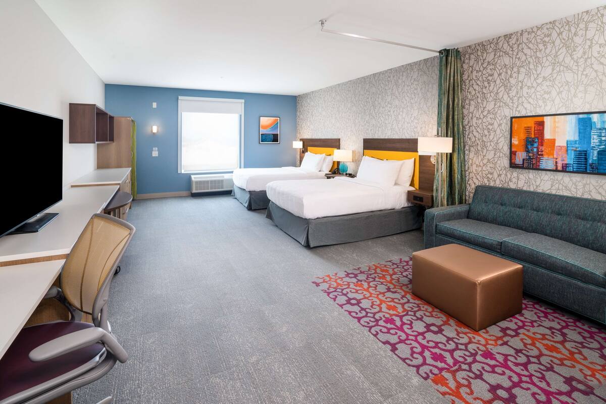 Habitación de hotel con dos camas, un escritorio, un televisor, un sofá y una alfombra estampada.